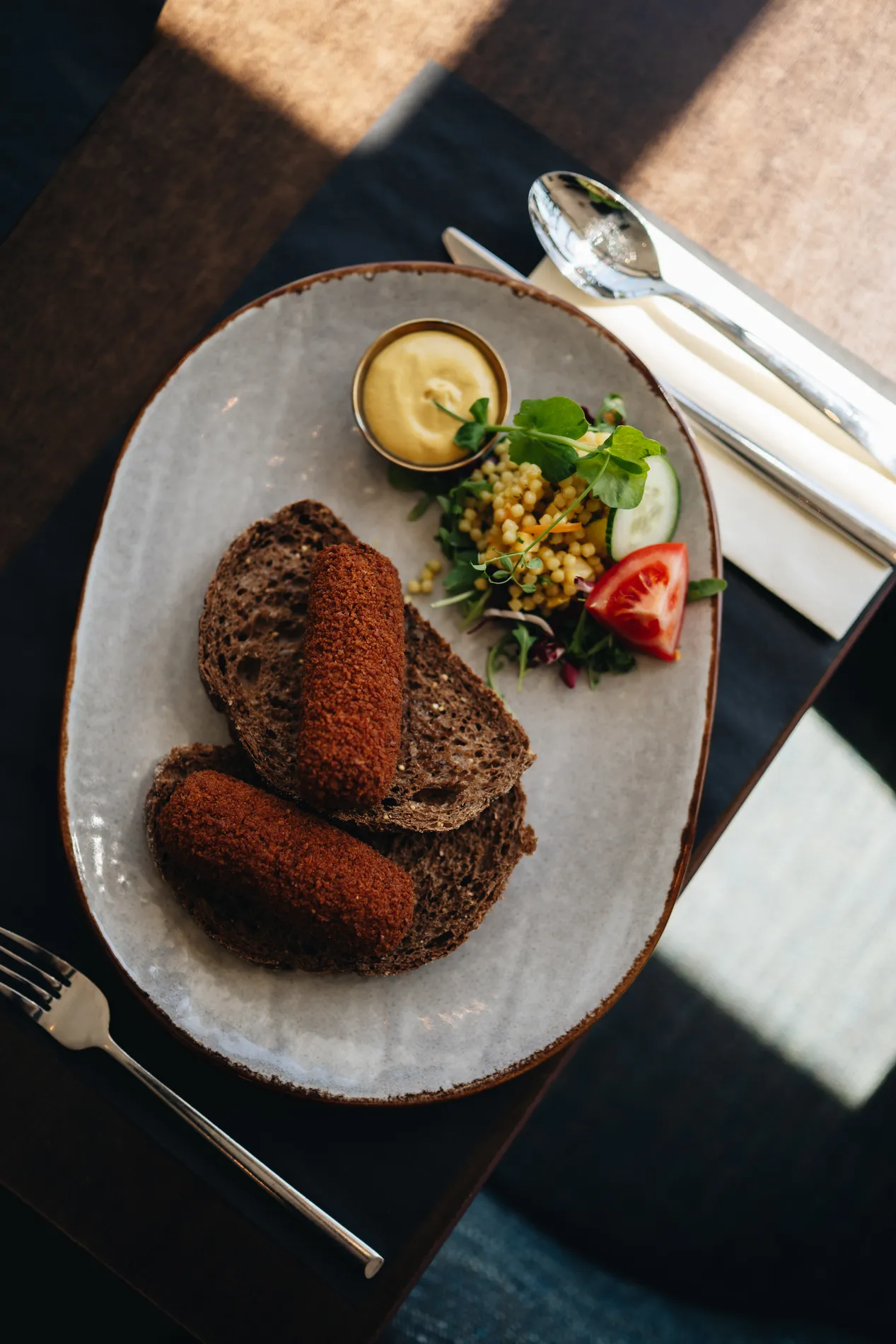 Lunchgerecht met twee ambachtelijke, bruine kroketten op dikke sneden donkerbruin brood, geserveerd met een schaaltje mosterd en een salade met parelcouscous.